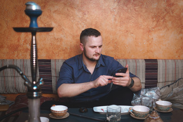 a man of European appearance, relaxes and relaxes in a hookah, Turkish style, oriental tea ceremony, uses the phone, smartphone, answers the call, uses the Internet