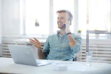 Portrait of handsome young man laughing cheerfully while speaking by phone sitting at desk in...