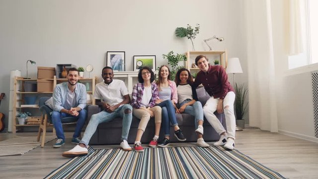 Portrait of young men and women happy friends running to sofa then sitting and looking at camera smiling and posing. Apartment, friendship and people concept.