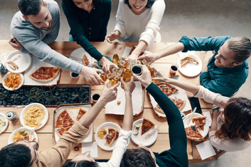 Fototapeta na wymiar To our friendship! Top view of young people in casual wear toasting each other and smiling while having a dinner party indoors