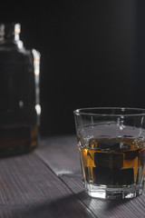 Vertical photo of a glass of whiskey and ice stones stands in the foreground on a wooden table in the background a fragment of a bottle