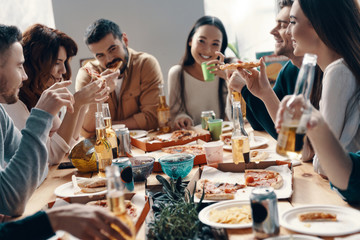 Totally carefree. Group of young people in casual wear eating pizza and smiling while having a dinner party indoors