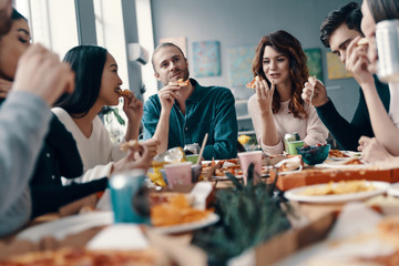 Great food and great people. Group of young people in casual wear eating pizza and smiling while having a dinner party indoors