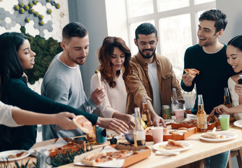 Enjoying hot pizza. Group of young people in casual wear eating pizza and smiling while having a dinner party indoors