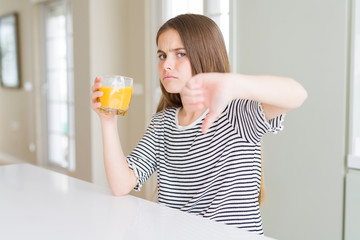 Beautiful young girl kid drinking a glass of fresh orange juice with angry face, negative sign showing dislike with thumbs down, rejection concept