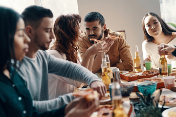 Sharing delicious meal. Group of young people in casual wear eating pizza and smiling while having a dinner party indoors