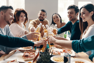 Toast to friends! Group of young people in casual wear toasting each other and smiling while having a dinner party indoors