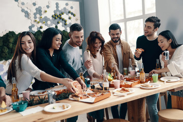 Delicious pizza. Group of young people in casual wear eating pizza and smiling while having a dinner party indoors