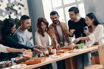 So hungry! Group of young people in casual wear eating pizza and smiling while having a dinner party indoors