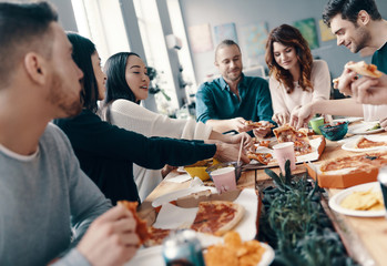 Feeling hungry. Group of young people in casual wear picking pizza and smiling while having a dinner party indoors