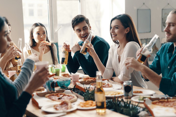 Relaxing evening. Group of young people in casual wear eating pizza and smiling while having a dinner party indoors
