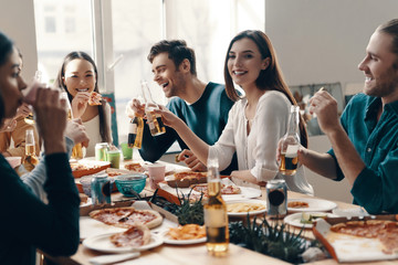 Having a great time. Group of young people in casual wear eating pizza and smiling while having a dinner party indoors