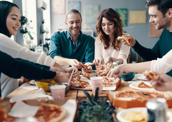 Pizza lovers. Group of young people in casual wear picking pizza and smiling while having a dinner party indoors