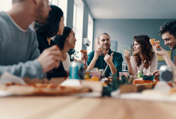 Life is better with friends. Group of young people in casual wear eating pizza and smiling while having a dinner party indoors