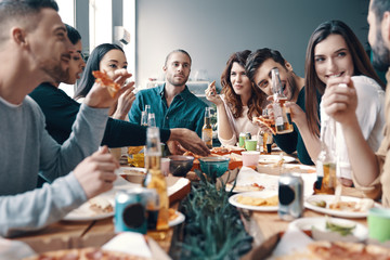 Meeting with best friends. Group of young people in casual wear eating pizza and smiling while having a dinner party indoors
