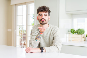 Fototapeta premium Young man wearing casual sweater sitting on white table looking at the camera blowing a kiss with hand on air being lovely and sexy. Love expression.