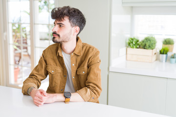 Fototapeta premium Young man wearing casual jacket sitting on white table looking to side, relax profile pose with natural face with confident smile.