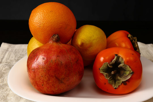 Close-up image. Still life with ripe tropical fruits. Bright orange oranges, yellow lemons, ripe and juicy persimmons and pomegranates lying on a white serving plate standing on a table with a dark ba