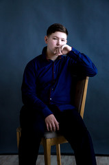 A complete teenager in a blue shirt is sitting on a wooden chair, against a dark background, business style.