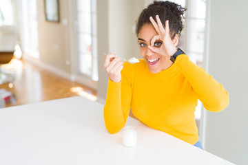 Young african american woman eating a healthy natural yogurt with happy face smiling doing ok sign with hand on eye looking through fingers