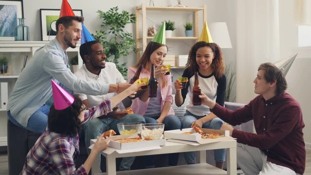 Happy young people wearing party hats are celebrating birthday eating and drinking at home together clinking bottles toasting. Celebration and youth concept.