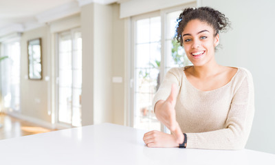 Beautiful young african american woman with afro hair sitting on table at home smiling friendly offering handshake as greeting and welcoming. Successful business.