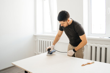 Young man working with sanding machine in the hands in a bright room polishes a wooden worktop. Concept repair at home.