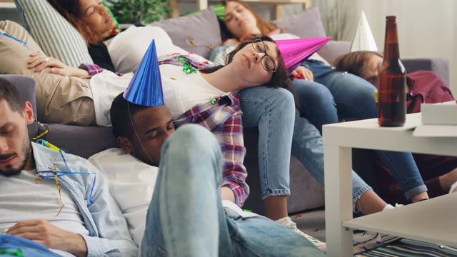 Exhausted girls and guys in bright hats are sleeping on floor and sofa after party in apartment. Youth lifestyle, celebrations and millennials concept.