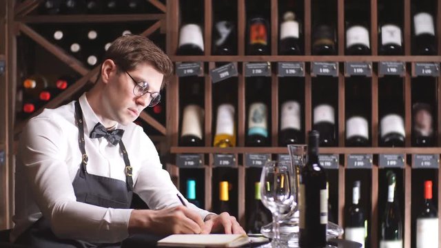 Experienced male wine steward doing stocktaking of wine at store, writing in notebook some notes in dark room with rows of racks