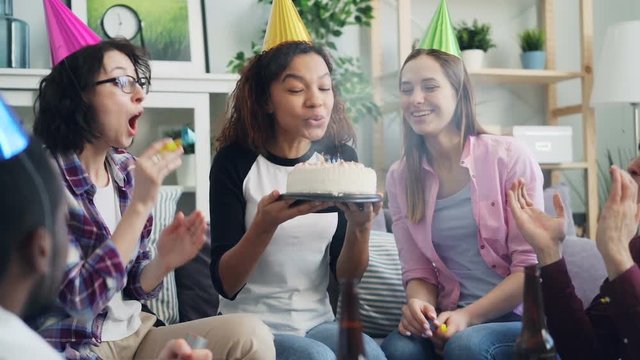 Slow motion of happy African American woman blowing candles on birthday cake celebrating with friends laughing having fun. Men and women blowing party whistles and throwing confetti.