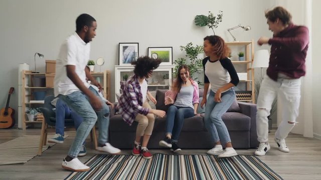 Slow motion portrait of joyful girls and guys running to sofa sitting smiling and looking at camera with happy faces. Apartment, cheerful youth and emotions concept.