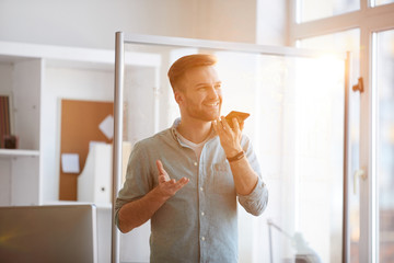 Waist up portrait of handsome young man  speaking by phone standing by window in sunlight, copy...