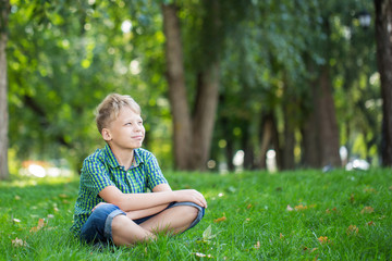 Cute white kid sitting on green grass at park. Horizontal color photography.