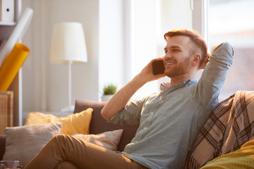 Portrait of handsome young man speaking by phone sitting on couch in sunlight, copy space
