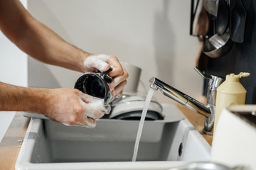 Homework, male hands with a sponge in the foam wash a dark mug in the kitchen sink with detergent.