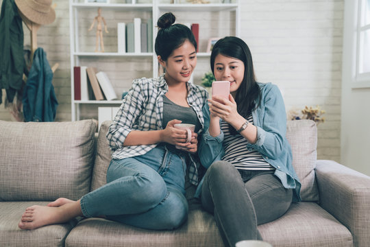 Young happy female asian watching online content in a smart phone sitting on sofa at home in living room. two girls roommates friends hold cup of tea showing cellphone screen with summer sale website
