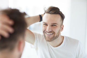 Portrait of handsome young man with lush hair and short stubble looking at his reflection in mirror and smiling, copy space
