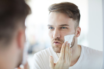 Head and shoulders portrait of handsome young man applying shaving cream  looking at his reflection in mirror, copy space