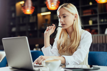 Young Caucasian blonde businesswoman dressed smart casual using laptop and holding pen while...