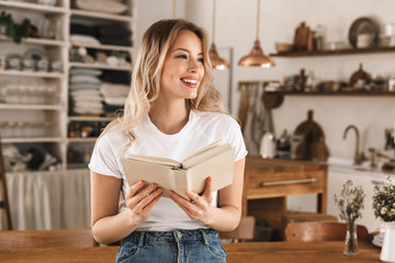Portrait of pretty blond woman reading book while standing in stylish wooden kitchen at home