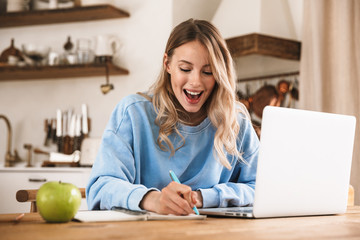 Portrait of beautiful blond woman 20s wearing casual sweatshirt working on laptop and writing down...
