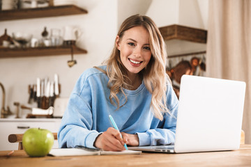 Portrait of smiling blond woman 20s wearing casual sweatshirt working on laptop and writing down notes at home - obrazy, fototapety, plakaty