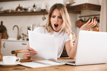 Image of stressed blond woman studying on laptop and writing down notes while sitting at wooden table in apartment