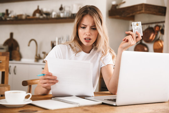 Image of serious blond woman holding plastic credit card while looking at paper documents in apartment