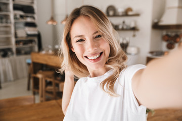 Image closeup of gorgeous blond woman looking at camera and taking selfie photo in living room