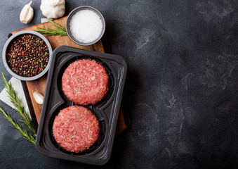 Fototapeta na wymiar Plastic tray with raw minced homemade beef burgers with spices and herbs. Top view and space for text on stone kitchen table background with tomatoes salt and pepper.