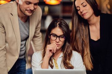 Charming buisnesswoman with brown hair and dressed elegant sitting at restaurant and looking at laptop. Next to her standing two colleagues and looking at laptop, too.