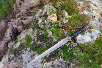 mysterious and magical photo of silver sword over the stone covered with moss in the England woods. Medieval period concept
