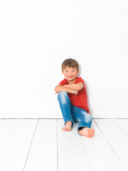 cute and blond boy with red shirt and blue jeans is posing on white wooden floor in front of white background