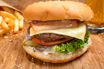 Big tasty cheeseburger on the wooden table with french fries. Delicious lunch with cheese, tomatoos and salad. Fast food with beer.